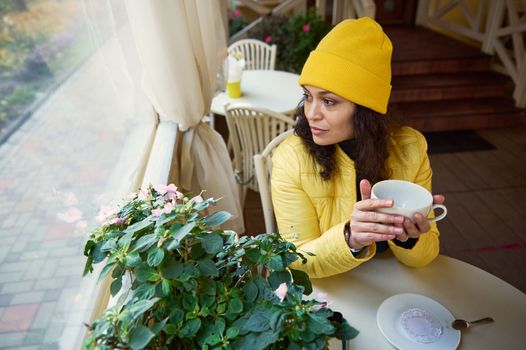 Millennial Latin American brunette woman in yellow hat and jacket, warms her hands with a cup of hot aroma coffee, looks out the window at the autumn nature while relaxing in a cozy coffee house
