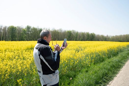 a middle-aged man motorcyclist stands in the middle of a field of flowering yellow rapeseed and photographs the landscape on a mobile phone using technology. High quality photo