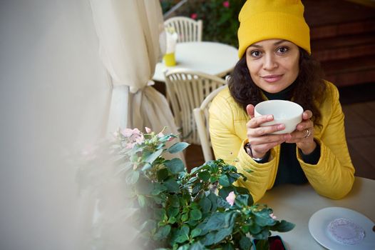 Beautiful dark-haired Latin American woman in yellow hat and jacket, sits at table with cup of hot drink in hands, smiles cutely looking at camera, chilling in a coffee shop with rustic vintage design