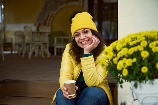 Delighted beautiful woman in bright yellow hat and jacket, sitting on a wooden doorstep with a takeaway coffee in a paper cup, next to potted colorful flowers,smiling a toothy smile looking at camera