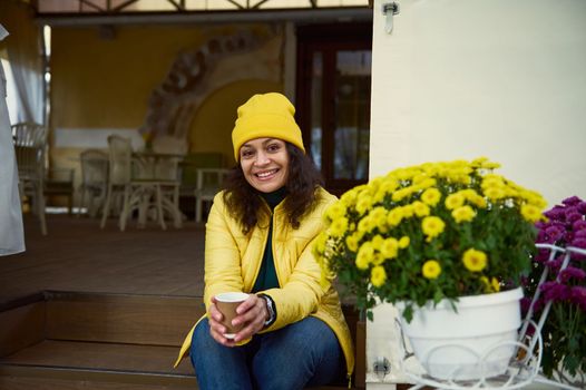 Attractive woman in yellow hat and jacket, smiling a toothy smile looking at camera, sitting on the wooden doorstep of a cozy coffee house, with a takeaway aroma coffee in a paper cup, on autumn day
