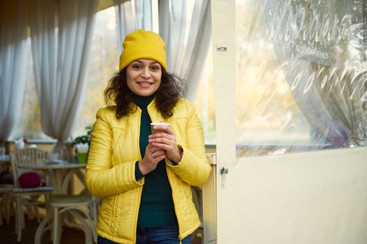 Attractive curly woman in green jacket, bright yellow hat and jacket warming hands with hot fragrant takeaway coffee in disposable paper cup, smiling beautiful toothy smile at camera