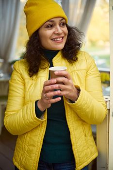 Close-up portrait of an attractive brunette multi-ethnic woman, in bright yellow hat and jacket, smiling a cheerful tothy smile looking aside, warming her hands with a takeaway hot coffee in paper cup