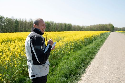 a middle-aged biker stands on the road in the middle of a field of flowering yellow rapeseed in belgium, looks into the distance and laughs. High quality photo