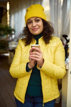 Confident dark-haired positive pretty woman in yellow hat and jacket, with a takeaway hot aroma coffee in a disposable paper cup, cutely smiling looking at camera. Portrait of diverse beautiful people