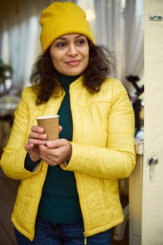 Waist-up portrait of a charming stunning positive multi-ethnic brunette woman, in yellow hat and jacket, smiling, looking thoughtfully aside, with takeaway hot coffee in paper cup in her hands