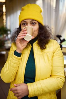 Close-up of a pretty woman in green sweater and bright yellow hat and jacket, taking a sip of hot aroma takeaway coffee in disposable paper cup