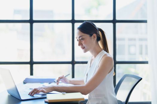 Portrait of a thoughtful Asian businesswoman looking at financial statements and making marketing plans using a computer on her desk.