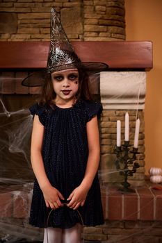 Charming little girl, a child witch in wizard hat, with a facial art makeup posing against a fireplace with spider web and candles on a candlestick. 31 October. Child celebrating Halloween. Autumn