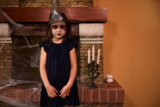 Mischievous mysterious Caucasian little child girl in witch costume and wizard hat, stands against a cobweb covered fireplace and candles, ready for Halloween party. Halloween trick or treat