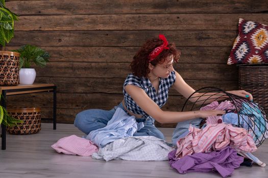 Attractive girl sorts clothes for washing. Short curly hair. Metal basket full of clothes prepared for washing. A wall of brown boards.