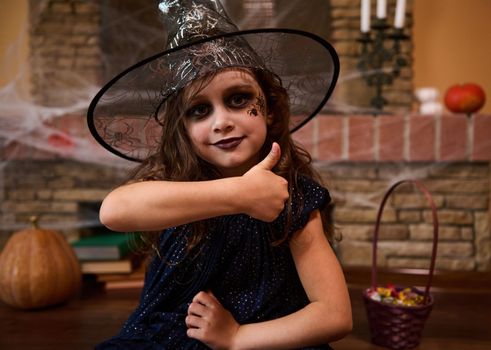 Close-up portrait of a gothic little child girl, with a spider painted on her cheek, wearing a wizard's hat and witch dress, shows thumb up, looking at camera. Halloween party. Enchantress. Sorceress