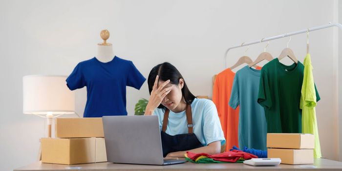 Woman seller SME retail store owner work hard worry stress in bad news on laptop at home office desk. Asian people crisis tailor job issue feel tired tough and upset in cash money loss or loan debt..
