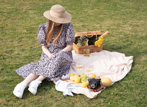 a young woman in a blue dress and straw hat is resting on a picnic with fruits, cheese plate and champagne, rest from worries and household chores parks and recreation areas,.High quality photo