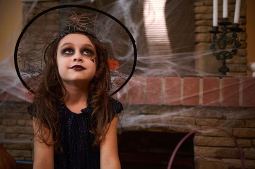 Mysterious little child girl with a spider on her cheek, in black dress and wizard's hat, sits against a cobweb covered fireplace with old candles on a vintage candlestick. Halloween trick and treat.