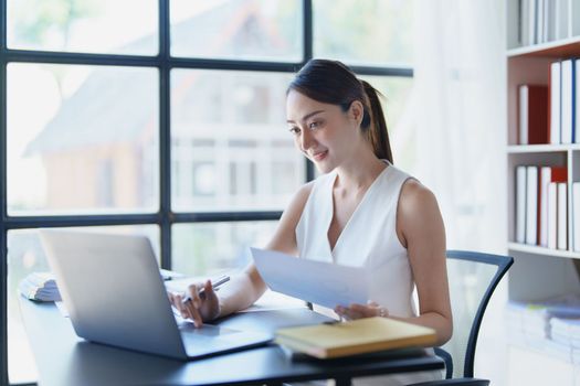 Portrait of a thoughtful Asian businesswoman looking at financial statements and making marketing plans using a computer on her desk.