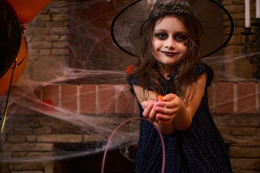 Caucasian cute little girl with a smoky eyes makeup, dressed as a sorceress with a wizard's hat, smiles looking at camera, holding a handful of candies, standing against a spider web covered fireplace