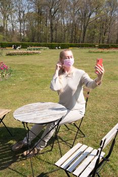 young woman in a beige jumper and a protective mask takes a selfie in the park against the backdrop of blooming tulips in early spring social media using a smartphone, High quality photo