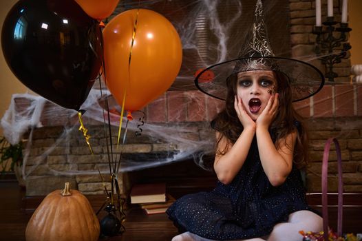 Charming Caucasian little girl, dressed in witch and wizard hat, sitting near orange black balloons and pumpkin, against a cobweb-covered fireplace. Halloween party. Gothic festivity. October 31