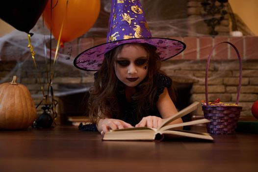 Adorable Caucasian child girl looking like a witch in a wizard hat, lying on the floor and studying a spell book, surrounded by a fireplace covered with cobwebs, a pumpkin, black and orange balloons