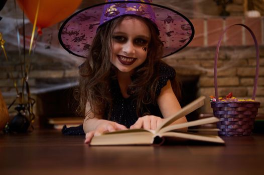 Pretty child, little Caucasian girl with face art makeup, wearing a wizard's hat, looking like a witch, posing with a sorcery book, smiling at the camera. Happy Halloween in festive gothic environment