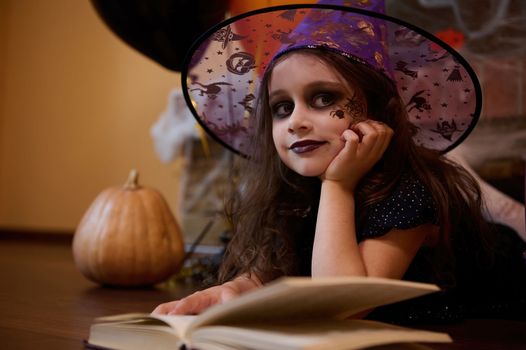 Adorable little child with a black spider painted on her cheek, looking like sorceress in wizard hat, smiles at camera while lying on the floor surrounded by a book of spells, pumpkin, Halloween decor