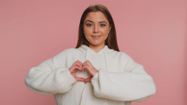 Woman in love. Smiling woman 20s in white sweater makes heart gesture demonstrates love sign expresses good feelings and sympathy. Young pretty adult girl isolated alone on pink studio background