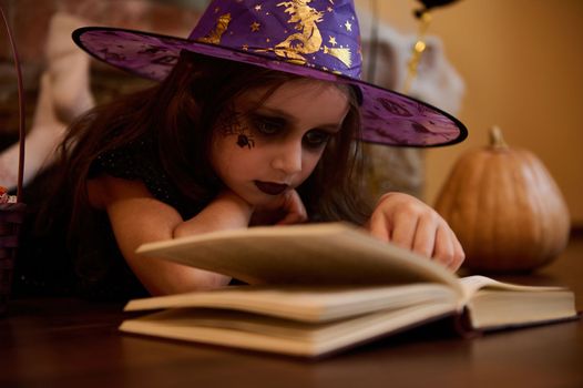 Close-up little girl with a face art make-up for Halloween, dressed as a witch, enchantress in wizard hat reading sorcery and spell magic book surrounded by pumpkins and festive gothic atmosphere