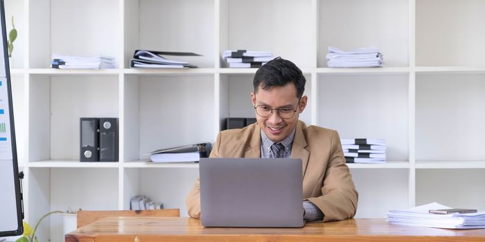 Happy young businessman in suit looking at laptop excited by good news online, lucky successful winner man standing at office desk raising hand in yes gesture celebrating business success win result..