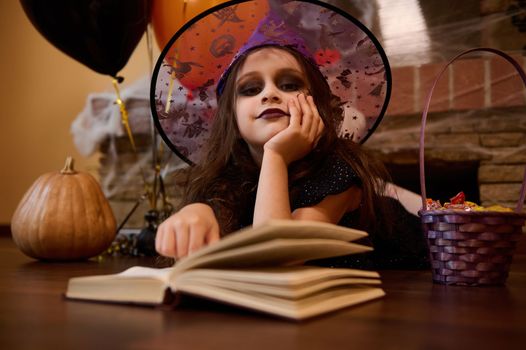 Adorable little child girl with face art make-up, in wizard hat, lying on the floor and reading book of spells, enchanter and sorcery, surrounded by pumpkins and a purple basket with Halloween treats