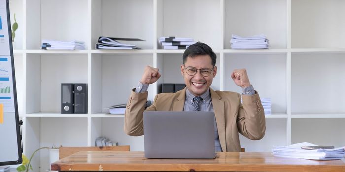Happy young businessman in suit looking at laptop excited by good news online, lucky successful winner man standing at office desk raising hand in yes gesture celebrating business success win result..
