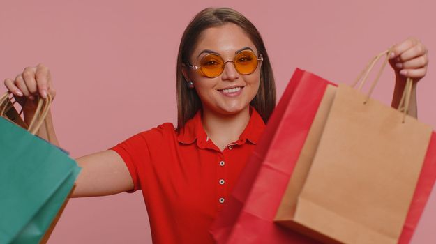 Happy tourist woman in red t-shirt showing shopping bags, advertising discounts, smiling looking amazed with low prices, shopping on Black Friday holidays. Young millennial girl on pink background