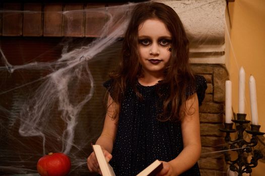 Caucasian gothic child with a spider paint on her cheek, little girl sorceress, witch, enchantress with a spell book, sits against a cobweb-covered fireplace and pumpkins, looking at camera. Halloween