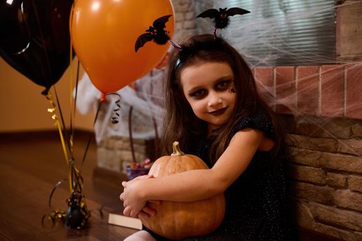 Portrait of an adorable European child girl, a little sorceress with a bat hoop, sitting by a fireplace covered with cobwebs and orange and black balloons, hugging a Halloween pumpkin. Jack-O-Lantern