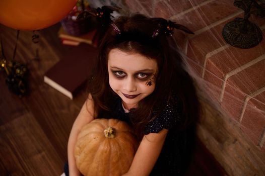 Top view Adorable gothic child, a little girl witch, enchantress, sorceress with face art makeup for Halloween party, hugging a pumpkin, smiles at camera, surrounded by a sorcery book and spider webs