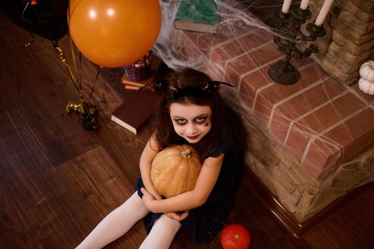 Top view of a little girl with gothic smoky eyes makeup, looks at camera and holds a Halloween pumpkin Jack-o-Lantern, sitting by a cobweb covered fireplace, candles, air balloons and spells books