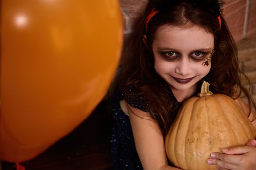 Close-up. Top view of a charming Caucasian little girl with face art makeup - a black spider painted on her cheek, looks like a witch, enchantress hugging pumpkin, smiles at camera. Happy Halloween