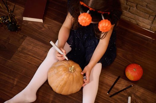 Top view of an adorable little girl in a black dress and pumpkins on a hoop, making a Jack-O-Lantern at home, painting a scary face on a pumpkin. Halloween party concept. October 31. autumn holiday