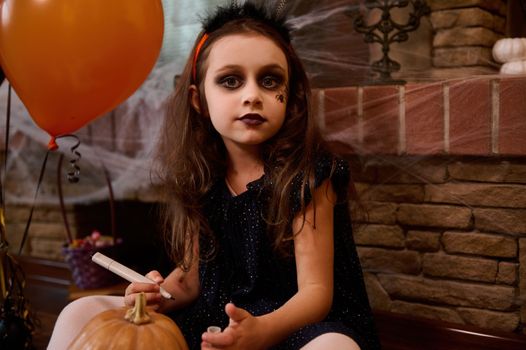 Beautiful little girl in a black dress, looking as a witch,making a Jack-O-Lantern at home, painting a scary face on a pumpkin. Halloween party concept. October 31. autumn holiday