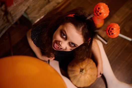 Top view of a charming Caucasian little girl with gothic smoky eyes makeup, looks at the camera and holds a Halloween pumpkin Jack-o-Lantern. Halloween treat and trick. October 31. Autumn festivity