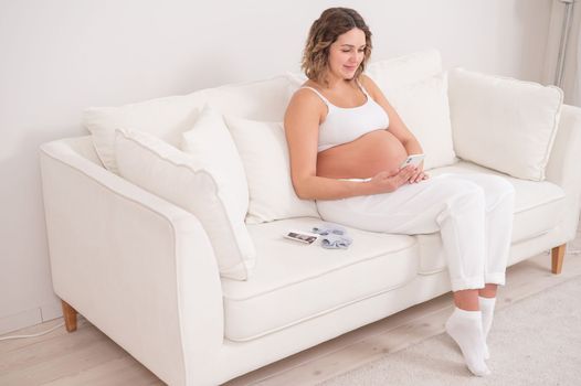 Pregnant woman sits on a white sofa and holds a smartphone. Fetal ultrasonic screening