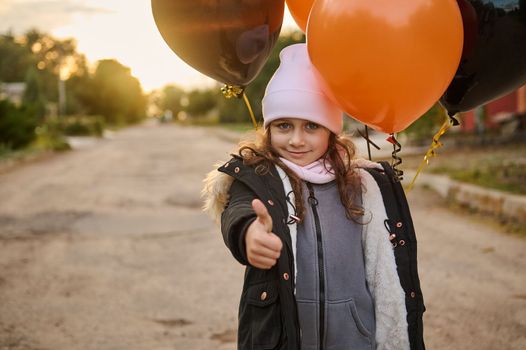Adorable beautiful Caucasian little girl with black and orange balloons walking down the street on an autumn day and showing a thumb up, looking at the camera. Halloween. October 31