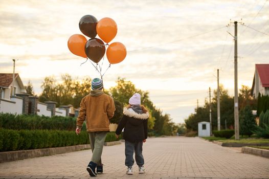 Rear view of elementary age kids, boy and girl or brother and sister, with orange black balloons, walking down the country street at sunset on Halloween. Autumn. Gothic festivity. October 31