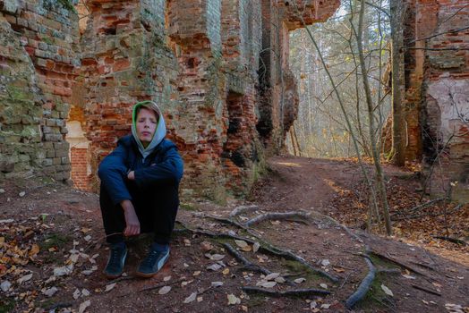 Sad Teen sit near the Brick Wall of the Old HouseA 12-15-year-old boy sits in a blue jacket near an old red brick wall. A teenager sits in ruins of brick walls.High quality photo