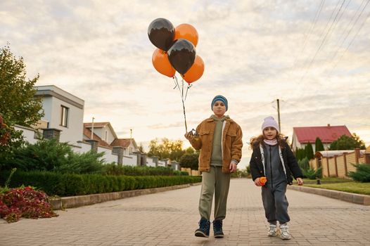 Adorable diverse Caucasian kids, boy and girl or brother and sister, with orange black balloons, walking down the country street at sunset on Halloween. Autumn. Gothic festivity. October 31