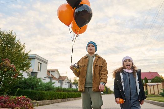 Handsome teenage boy and his younger sister, walk down the street with black and orange air balloons, celebrating Halloween, standing on the village road at beautiful sunset background