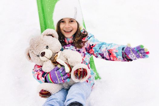 Portrait of child with toy teddy bear sits on a sled and looks at the winter snowy mountains.Winter vacantion. Christmas celebration and winter holidays. Winter fun and outdoor activities with kids.