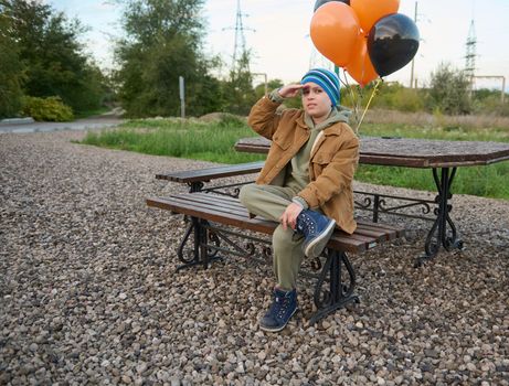 Handsome Caucasian teenage boy with black and orange air balloons, sitting on a wooden bank and looking forward. Happy Halloween. October 31. Gothic festivity