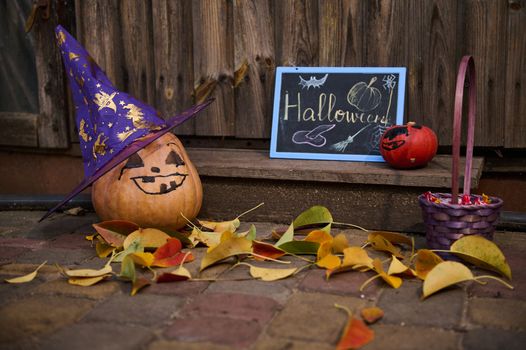 Halloween treat and trick. Closeup of a whole orange pumpkin with a painted scary face in a purple wizard's hat, next to a blackboard and fallen autumnal dry colorful leaves on the wooden doorstep