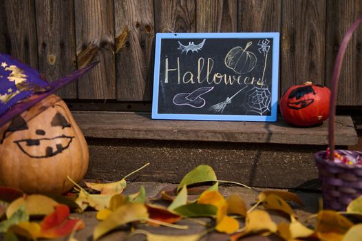 Selective focus. A blackboard with the inscription Halloween stands next to pumpkins on a wooden threshold with fallen yellow and red leaves in autumn. Porch or backyard decoration for Halloween